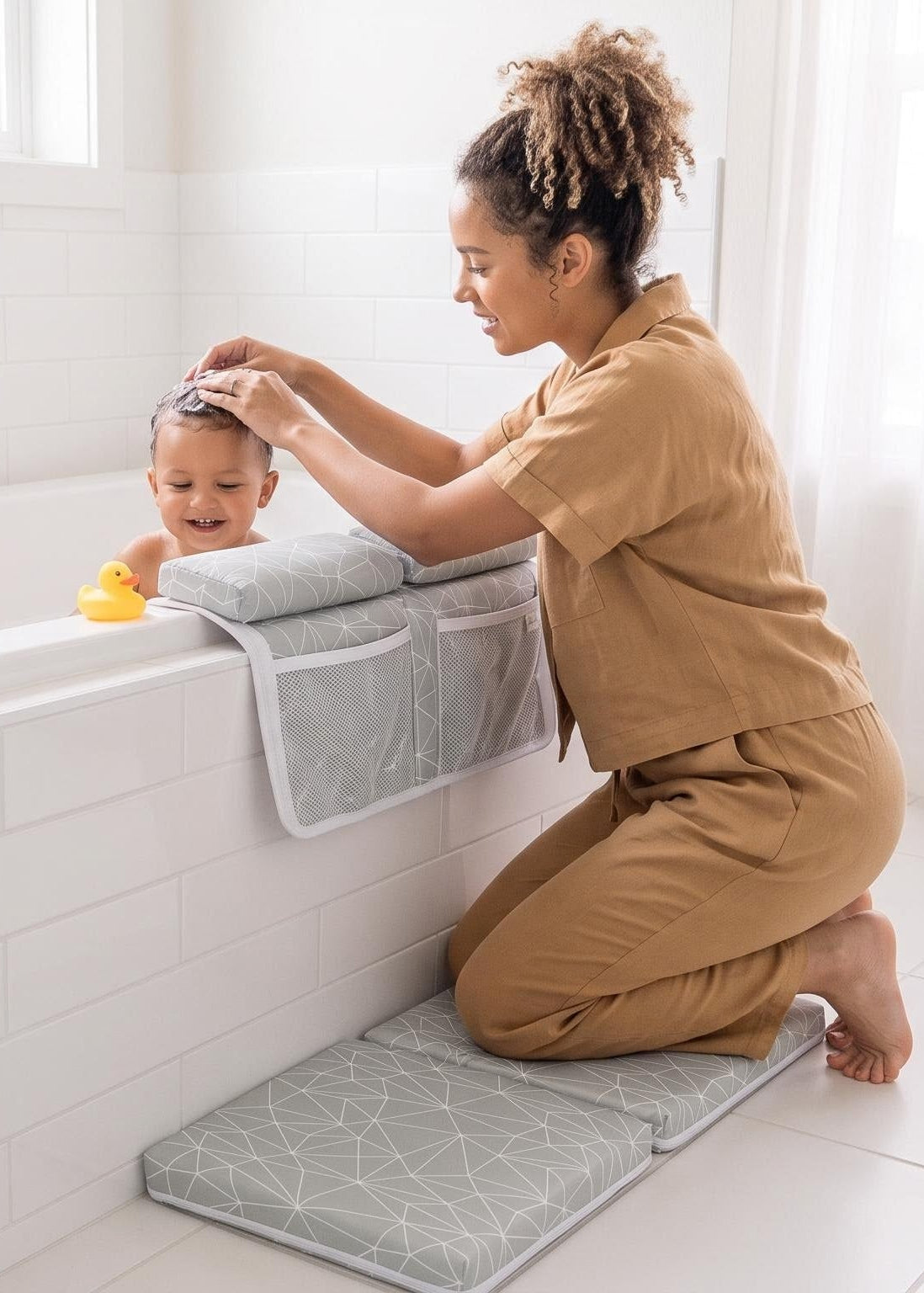 A woman in a tan scrub-style uniform kneels beside a white bathtub in a bright bathroom, holding a baby on a gray padded support pad. A folded towel rests on the tub edge, with a window and white curtains in the background.