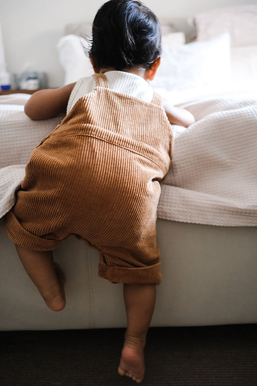 baby climbing up  the bed wearing corduroy overalls